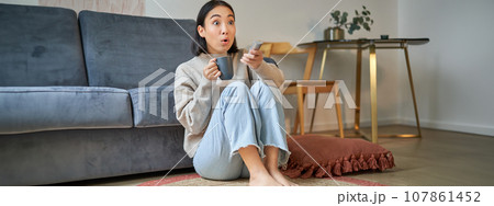 Portrait of girl watching television at home, sits on floor near sofa, holds remote and changes channels Portrait of girl watching television at home, sits on floor near sofa, holds remote and changes channels 107861452