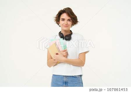 Image of brunette woman in headphones, standing with documents. Student with notes and workbooks posing against white background 107861453