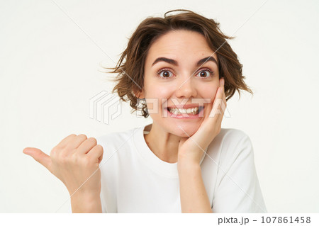 Image of excited brunette woman with short haircut, pointing left, smiling and looking happy, showing you advertisement, standing over white background Image of excited brunette woman with short haircut, pointing left, smiling and looking happy, showing you advertisement, standing over white background 107861458