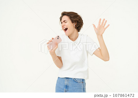 Portrait of happy young woman singing in mobile speakerphone, listening music in wireless headphonse, playing smartphone karaoke game, standing over white background Portrait of happy young woman singing in mobile speakerphone, listening music in wireless headphonse, playing smartphone karaoke game, standing over white background 107861472