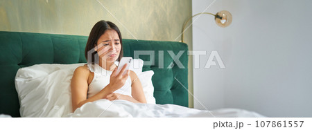 Portrait of concerned asian woman holding mobile phone, receive bad phone call, looking worried and upset, having difficult telephone conversation while lying in bed Portrait of concerned asian woman holding mobile phone, receive bad phone call, looking worried and upset, having difficult telephone conversation while lying in bed 107861557