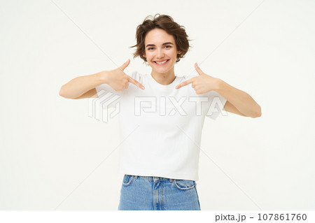 Image of confident, good-looking young woman, smiling and pointing at herself, standing over white studio background 107861760