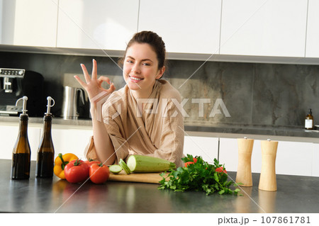 Portrait of smiling brunette girl making vegan dinner, showing okay sign, recommending healthy food, chopping vegetables in kitchen for salad or for vegan breakfast 107861781