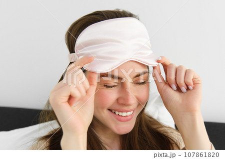 Close up portrait of gorgeous smiling woman with sleeping mask on forehead, waking up in morning, relaxing in bed, laughing and looking happy, resting on vacation in hotel room 107861820