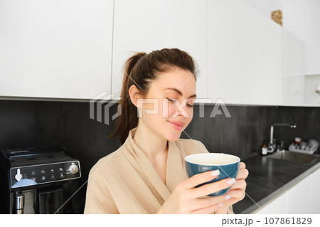 Portrait of good-looking young woman starting her day with cup of coffee, standing in the kitchen and drinking cappuccino from big mug, enjoying favourite drink in the morning 107861829