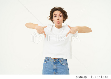 Portrait of amazed girl, pointing fingers down, showing something and looking surprised, standing over white studio background 107861838