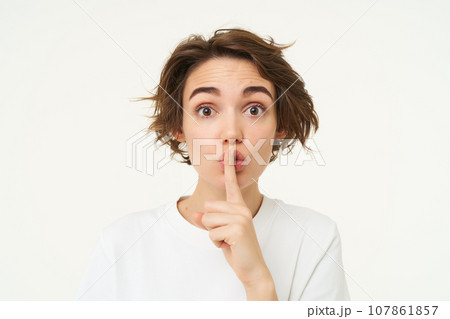 Close up shot of cute girl, press finger to lips, makes shhh shush noise, keeps secret, stands over white background 107861857