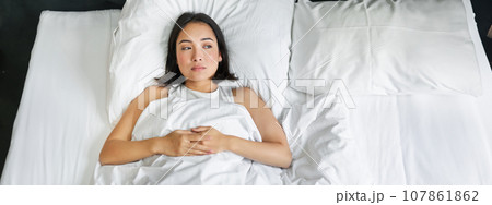 Portrait of asian girl lying alone in bed with white sheets bedding, looking thoughtful. Woman in her bedroom thinking of smth and gazing at window Portrait of asian girl lying alone in bed with white sheets bedding, looking thoughtful. Woman in her bedroom thinking of smth and gazing at window 107861862