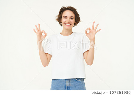 Portrait of young woman, showing okay, ok gestures and smiling, giving positive feedback, recommending, standing over white background 107861986