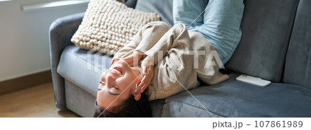 Portrait of happy young asian woman feeling upbeat, lying on sofa and listening music in wireless headphones, smiling pleased 107861989