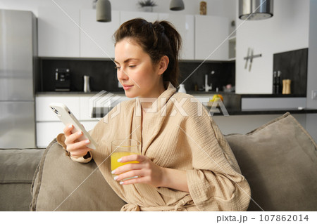 Lifestyle and morning concept. Beautiful woman sitting on sofa and resting, reading news on smartphone, drinking orange juice at home 107862014