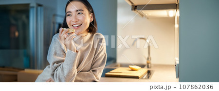 Portrait of smiling young happy woman staying at home, standing in kitchen and eating toast, looking aside 107862036