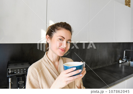 Portrait of good-looking young woman, drinking coffee in the kitchen, enjoying her morning routine and smiling at the camera 107862045