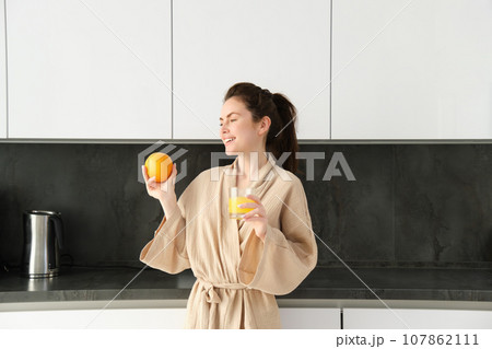 Portrait of attractive young woman, posing with an orange, drinking fresh juice at home in kitchen, starting her morning with healthy drink 107862111
