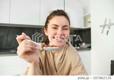 Portrait of smiling beautiful woman, feeding you spoon of cereals with milk, eating breakfast and looking happy, enjoying her morning, wearing cosy bathrobe Portrait of smiling beautiful woman, feeding you spoon of cereals with milk, eating breakfast and looking happy, enjoying her morning, wearing cosy bathrobe 107862118