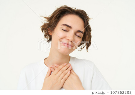 Close up portrait of young woman feeling warmth and love, holds hands on heart and smiles, stands over white studio background Close up portrait of young woman feeling warmth and love, holds hands on heart and smiles, stands over white studio background 107862202