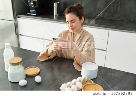 Attractive young cheerful girl baking at the kitchen, making dough, holding recipe book, having ideas Attractive young cheerful girl baking at the kitchen, making dough, holding recipe book, having ideas 107862298