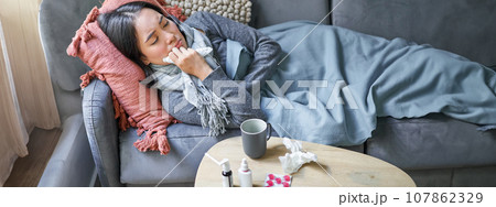 Sick sad korean woman lying on sofa, feeling unwell, catching cold, flu and temperature, looking upset, taking medication Sick sad korean woman lying on sofa, feeling unwell, catching cold, flu and temperature, looking upset, taking medication 107862329