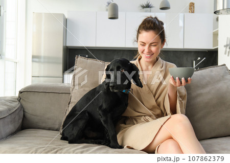 Happy young woman in bathrobe, enjoys spending time with her dog at home, eating cereals, having breakfast with puppy, sitting on sofa 107862379