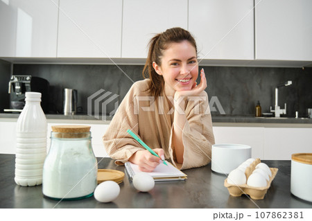 Attractive young cheerful girl baking at the kitchen, making dough, holding recipe book, having ideas 107862381