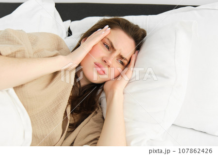 Close up portrait of sick woman lying in her bed and feeling unwell, touching head, has headache, looking frustrated from migraine, wearing pyjamas Close up portrait of sick woman lying in her bed and feeling unwell, touching head, has headache, looking frustrated from migraine, wearing pyjamas 107862426