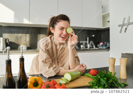 Portrait of brunette woman, wife cooking at home, making dinner, posing near chopping board in kitchen with vegetables, holding zucchini 107862521