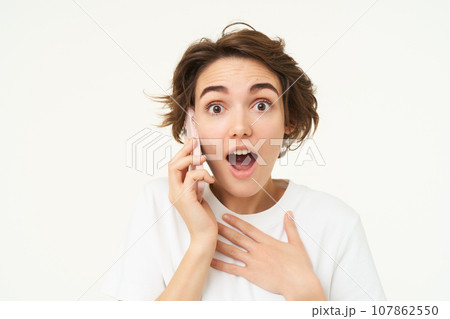 Portrait of woman with surprised face, answers phone call and looks excited, amazed by big news, stands over white background 107862550