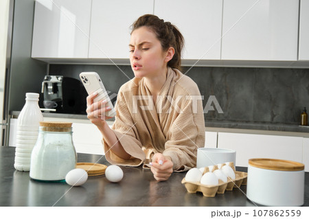 Attractive young cheerful girl baking at the kitchen, making dough, holding recipe book, having ideas Attractive young cheerful girl baking at the kitchen, making dough, holding recipe book, having ideas 107862559