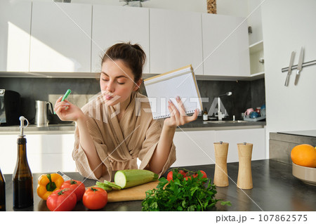Portrait of modern young woman cooking, making grocery list, reading recipe and making meal, salad in the kitchen, looking at vegetables on chopping board Portrait of modern young woman cooking, making grocery list, reading recipe and making meal, salad in the kitchen, looking at vegetables on chopping board 107862575
