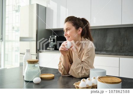 Attractive young cheerful girl baking at the kitchen, making dough, holding recipe book, having ideas 107862588