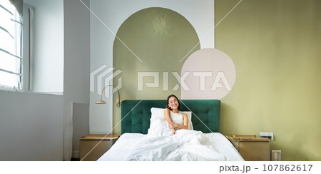 Vertical shot of beautiful asian girl lying in bedroom with sophisticated interrior, smiling and looking happy in morning, spending time in bed Vertical shot of beautiful asian girl lying in bedroom with sophisticated interrior, smiling and looking happy in morning, spending time in bed 107862617