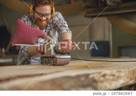 Close up of smiling bearded male wood worker using electric sander for wood. Carpentry workshop 107862667