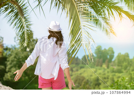 Silhouette of a woman from behind against the backdrop of tropical palm trees and bright sun. Summer vacation in tropical countries. 107864994