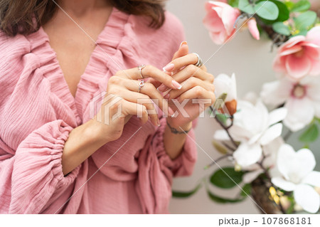 Close shot of romantic cute french gel polish manicure nails. Woman in pink summer dress with flowers around. Rings on fingers. Outdoor shot. 107868181