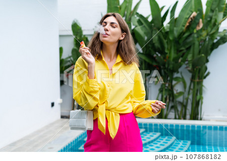 Stylish fit fashion women in bright pink pants holding bag trendy posing at luxury tropical villa by pool outdoor natural day light smoking vape pod enjoying puffs 107868182
