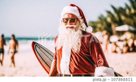 A modern tanned older man in Santa Claus costume walks along the beach with surfboard for swimming. Winter holidays 107868811