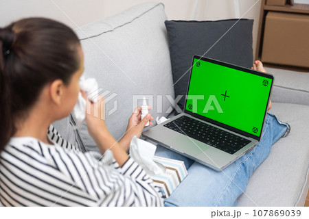 Young woman wipes nose with handkerchief talking to therapist on online call on laptop lying on sofa patient with influenza receives support of doctor remotely from home side view 107869039