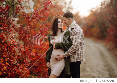 Happy and young pregnant couple in autumn park. People, lifestyle, relaxation and vacations concept. Happy and young pregnant couple in autumn park. People, lifestyle, relaxation and vacations concept. 107869220