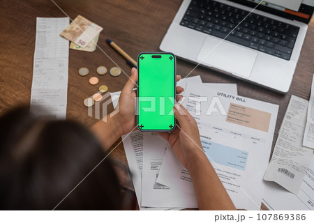 Mobile Financial Control: A woman's hand holds a mobile phone with a chroma key screen on a wooden desk, accompanied by utility bills, cash, and an online banking app.  107869386