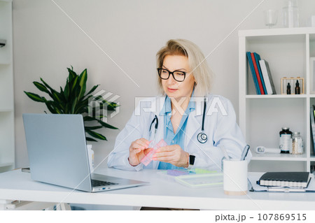 Friendly female middle aged doctor shows jar of pills and pill organizer to laptop in clinic office. Video call online, treatment remote, pharmacy, supplement advice, medical healthcare. Telemedicine 107869515