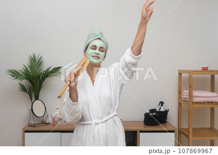 Bathrobe Karaoke: A joyful lady, clay mask on her face, sings into a hairbrush mic, dancing at home with a towel on her head, having a blast. Bathrobe Karaoke: A joyful lady, clay mask on her face, sings into a hairbrush mic, dancing at home with a towel on her head, having a blast. 107869967