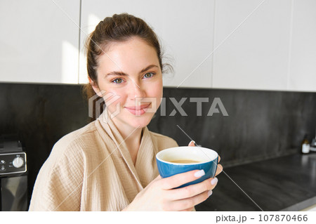 Good-looking young woman with cup of coffee, posing in the kitchen, smiling at camera, enjoys her morning at home, wearing cosy bathrobe 107870466