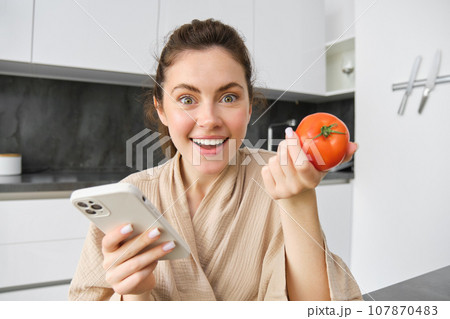 Close up portrait of beautiful young woman in the kitchen, holding tomato and smartphone, buys fresh vegetables online, orders groceries delivery on mobile phone application 107870483