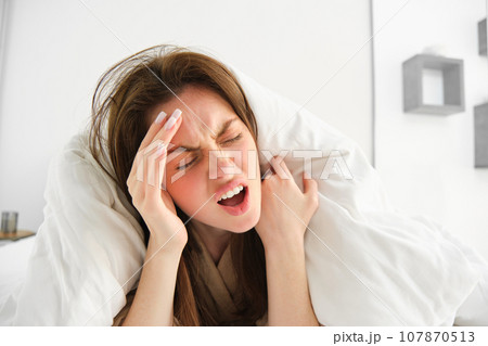 Woman suffering headache in morning, lying in bed under blankets, screaming from pain in head Woman suffering headache in morning, lying in bed under blankets, screaming from pain in head 107870513
