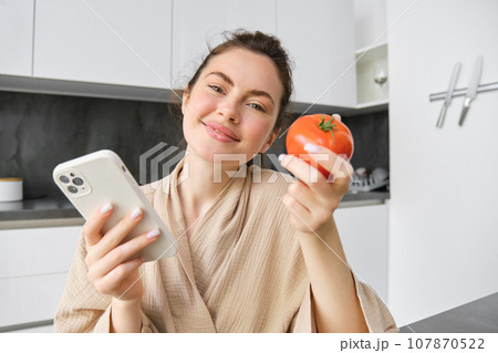 Close up portrait of beautiful smiling woman, holding fresh tomato, sitting in kitchen with smartphone, orders vegetables online, using application to buy groceries, using mobile phone Close up portrait of beautiful smiling woman, holding fresh tomato, sitting in kitchen with smartphone, orders vegetables online, using application to buy groceries, using mobile phone 107870522