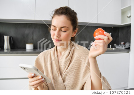 Close up portrait of beautiful smiling woman, holding fresh tomato, sitting in kitchen with smartphone, orders vegetables online, using application to buy groceries, using mobile phone 107870523