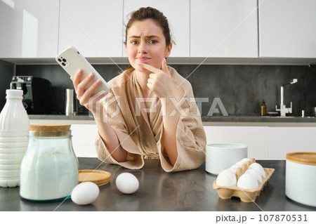 Attractive young cheerful girl baking at the kitchen, making dough, holding recipe book, having ideas Attractive young cheerful girl baking at the kitchen, making dough, holding recipe book, having ideas 107870531