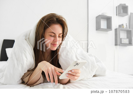 Woman in morning lying in bed covered in blanket, looking at smartphone, using mobile phone app Woman in morning lying in bed covered in blanket, looking at smartphone, using mobile phone app 107870583