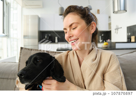 Close up portrait of young woman sitting on sofa and hugging adorable black dog, spending time with her puppy friend at home Close up portrait of young woman sitting on sofa and hugging adorable black dog, spending time with her puppy friend at home 107870897