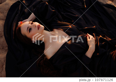 A woman in a black dress with a long train sits on a sand dune. 107871126